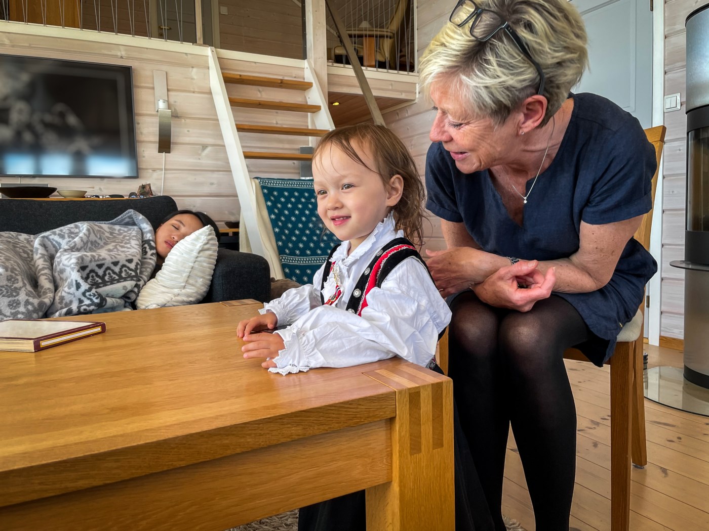 Bestemor og barnebarn har en varm samtale ved kjøkkenbordet, mens en annen voksen hviler på sofaen i bakgrunnen. Et øyeblikk av tillit og nærhet i en norsk stue.